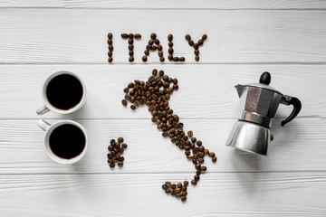 Map of the Italy made of roasted coffee beans laying on white wooden textured background with two coffee cups and coffee maker. Space for text