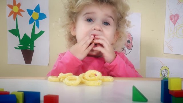 little girl eating corn circle sitting near her table toys and drawings