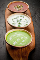 Selection of cold refreshing summer soups: tomato gazpacho, tarator, cream soup of green peas on a cutting board on a black concrete table close view copy space