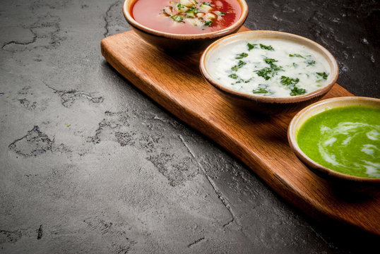 Selection of cold refreshing summer soups: tomato gazpacho, tarator, cream soup of green peas on a cutting board on a black concrete table close view copy space