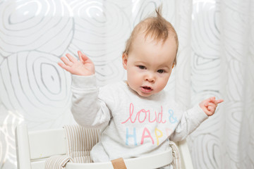 Eleven months old baby girl sitting in a stool making funny faces into the camera, raising her both hands in the air