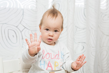 Fototapeta premium Eleven months old baby girl sitting in a stool making funny faces into the camera, raising her both hands in the air