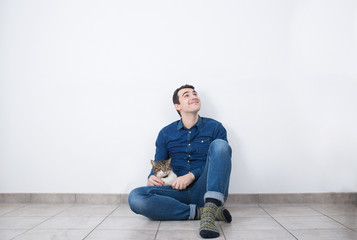 Young smiling man looking up sitting on the floor of new house with his cat.Property concept