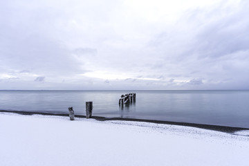 Tranquil sea with broken pier posts leading into the water, snow on winter beach