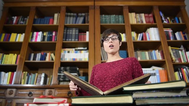 Young pretty female student with big library in background reading brown old book and suddenly move up face with happy expression