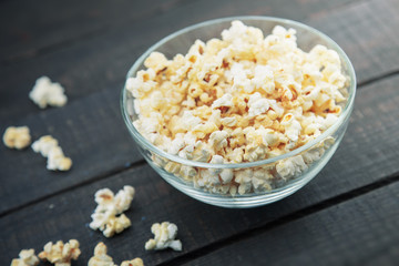 Glass bowl with freshly popped popcorn with salt on dark wooden background. Soft focus.
