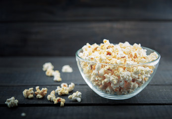 Glass bowl with freshly popped popcorn with salt on dark wooden background. Soft focus.