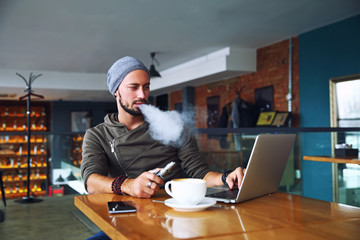 Young handsome hipster man with beard sitting in cafe with a cup of coffee, vaping and releases a cloud of vapor. Working at laptop and having a little break. With copy space. - 138628412