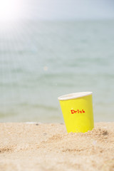 Yellow paper glass beverage on the sand behind beach with sunlight.