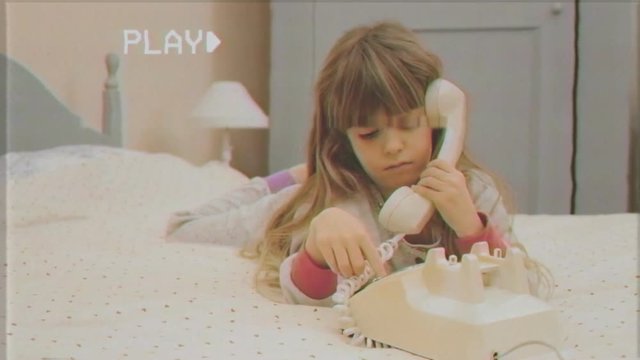 Fake VHS shot: a cute little girl lying on her bed, making a call with a vintage rotary phone.
