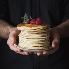 Stack of pancakes with raspberries in male hands, closeup view, selective focus. Square crop
