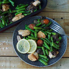 Square photo of hot salad with green beans, baked cherry tomatoes, chicken breast, sliced mushrooms, sesame seeds and lemon served in a grey plate with metal fork on wooden board