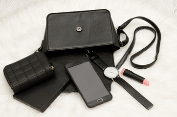 Open black bag with dropped things, notebook, mobile phone, watch, purse and lipstick. The white fur on background, top view. fashion concept