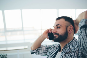 Side view of young bearded man,dressed incasual wear,sitting at round wooden table in cafe with modern interior and is holding smartphone.