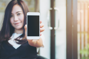 Mockup image of a beautiful woman holding and showing white mobile phone with blank black screen and smiley face in modern cafe