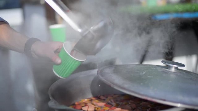 man cooking in large cauldrons and pours into glasses of mulled wine, outdoors, party, fair.