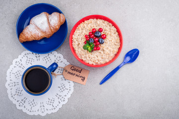 Coffee cup, oatmeal with berries, croissant and note good morning on grey background. Healthy breakfast concept. Top view. Copy space