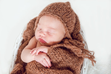 A newborn baby in a white wicker basket covered with a blanket knitted brown beanie sleep.