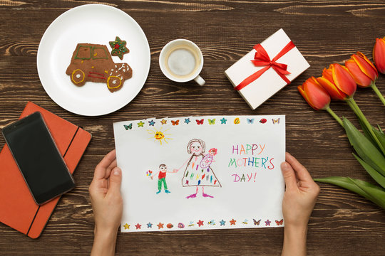 Happy Mothers Day Card Made By A Child. Mother's Hands Hold A Card A Gift From The Child And A Coffee Cup On A Wooden Background Of A Table.