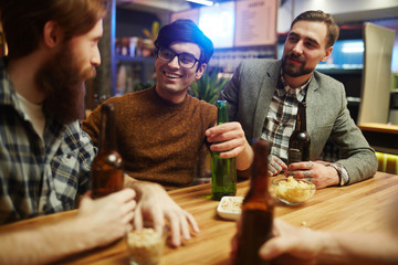 Happy young men with drinks sitting by table in pub and having talk