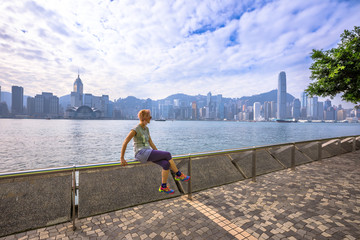 Naklejka premium Caucasian woman with urban background Hong Kong skyline. Female fitness athlete after a workout on Tsim Sha Tsui Promenade and Avenue of Stars in Victoria Harbour, Kowloon. Healthy lifestyle concept.