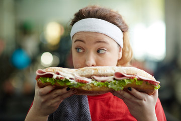 Closeup portrait of cute overweight woman holding big fattening sandwich in front of her face, eating it in secret and looking around cautiously, covered in smudges of mayo