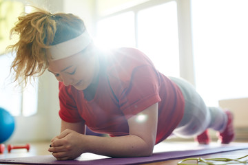 Portrait of young obese woman working out on yoga mat in fitness studio: holding plank exercise...