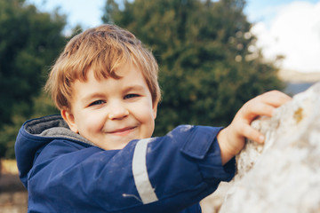 Close-up portrait 6 year old boy wearing jacket, happy smile on his face, blue eyes, blonde hair, rocks in nature