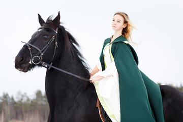 Young girl in white dress and green cape riding black thoroughbred horse in winter. Historical image