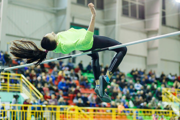 Young professional sportswoman jumping over bar in high jump competition against crowded grandstand.