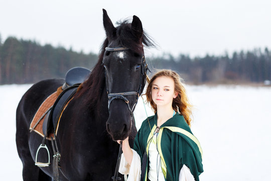 Young girl in white dress and green cape with black thoroughbred horse on winter field. Historical image