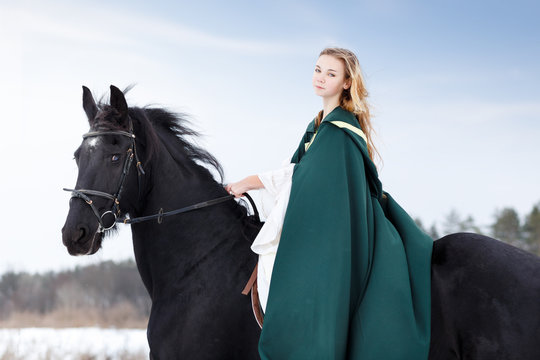 Young girl in white dress and green cape riding black thoroughbred horse in winter. Historical image