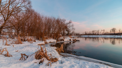 Beautiful evening winter landscape. February thaw. Coast of the river with the ice edge and the vegetation under the snow