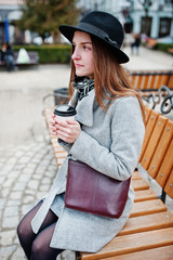 Fototapeta premium Young model girl in a gray coat and black hat with leather handbag on shoulders sitting on bench with plastic cup of coffee at street of city.