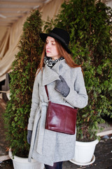 Young model girl in gray coat and black hat with leather handbag on shoulders posed against white cedar at street of city.