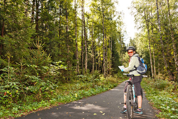 Obraz premium Female tourist cyclist standing on road in forest with map, looking to camera and smiling.