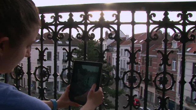 Girl sitting at terrace with wrought iron fencing and taking a photos on tablet pc, back view dolly shot