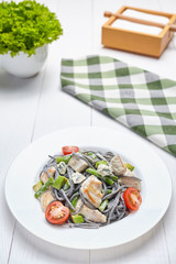 Black pasta meal with belsm sauce and slices of grilled meat and vegetables on a white plate. White background