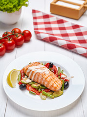 Salmon steak grilled with herbs and lemon, spicy white sauce and grilled vegetables on a white plate.