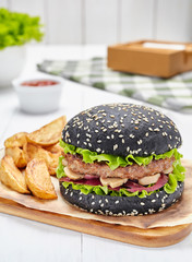 Black burger with beef, onions, tomatoes, lettuce and mushrooms, served with potato fries on a wooden board.