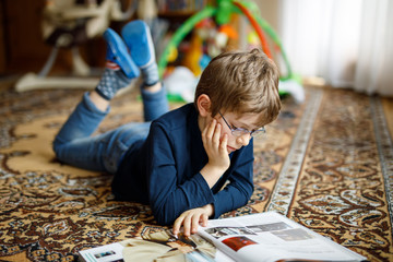 Little blond kid boy with eyeglasses reading book on floor at home