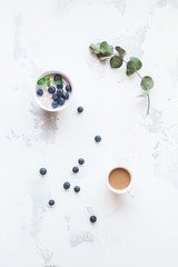 Cup of coffee, blueberry yogurt with muesli and eucalyptus branch on white background. Flat lay, top view