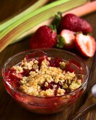 Baked strawberry and rhubarb crumble in glass bowl with fresh raw ingredients in the back,...