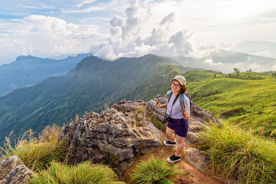 Hiker asian teen girl happy with camera backpack cap and eyeglass looking beautiful landscape nature of mountain range and colourful sky at sunset on Phu Chi Fa Forest Park, Chiang Rai, Thailand