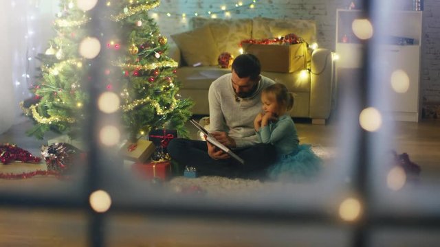 Looking Through the Window. Sitting Under Christmas Three with Gifts Under It Father Reads Book to His Daughter. Shot on RED Cinema Camera 4K (UHD).