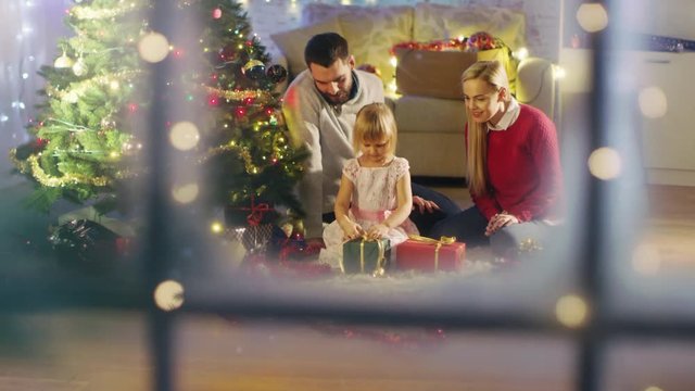 Looking Through Snowy Window. Happy Father, Mother and Daughter Sitting Under Christmas Tree. Daughter Opens Her Presents.Shot on RED Cinema Camera 4K (UHD).