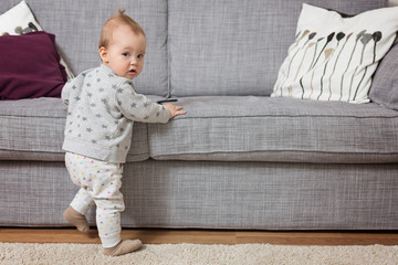 Eleven months old baby girl standing by the sofa, holding it with her tiny hands