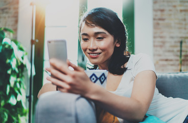 Young smiling Asian woman spending rest time at home, holding ceramic cup at hand and using smartphone for video conversation with friends.Blurred background, flares effect.