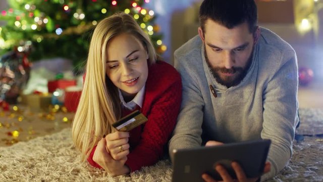 Happy Couple Lies On the Carpet Under Christmas Tree, Woman Holds Credit Card and Man Buys Her Presents on Tablet Computer. 
