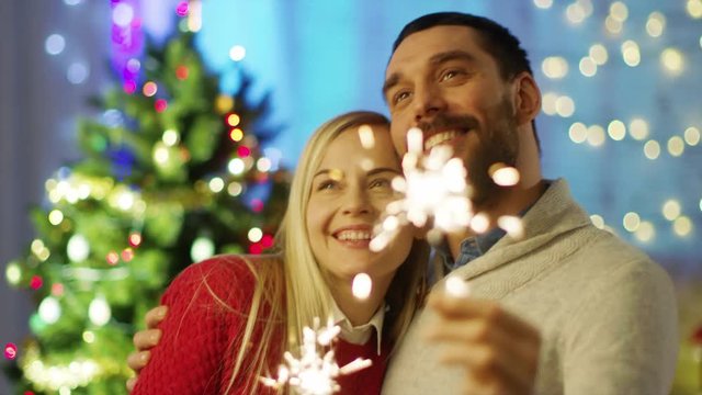 Happy Couple Light Sparklers and Smile. In the Background Christmas Tree and Room Decorated with Lights. Shot on RED Cinema Camera 4K (UHD).